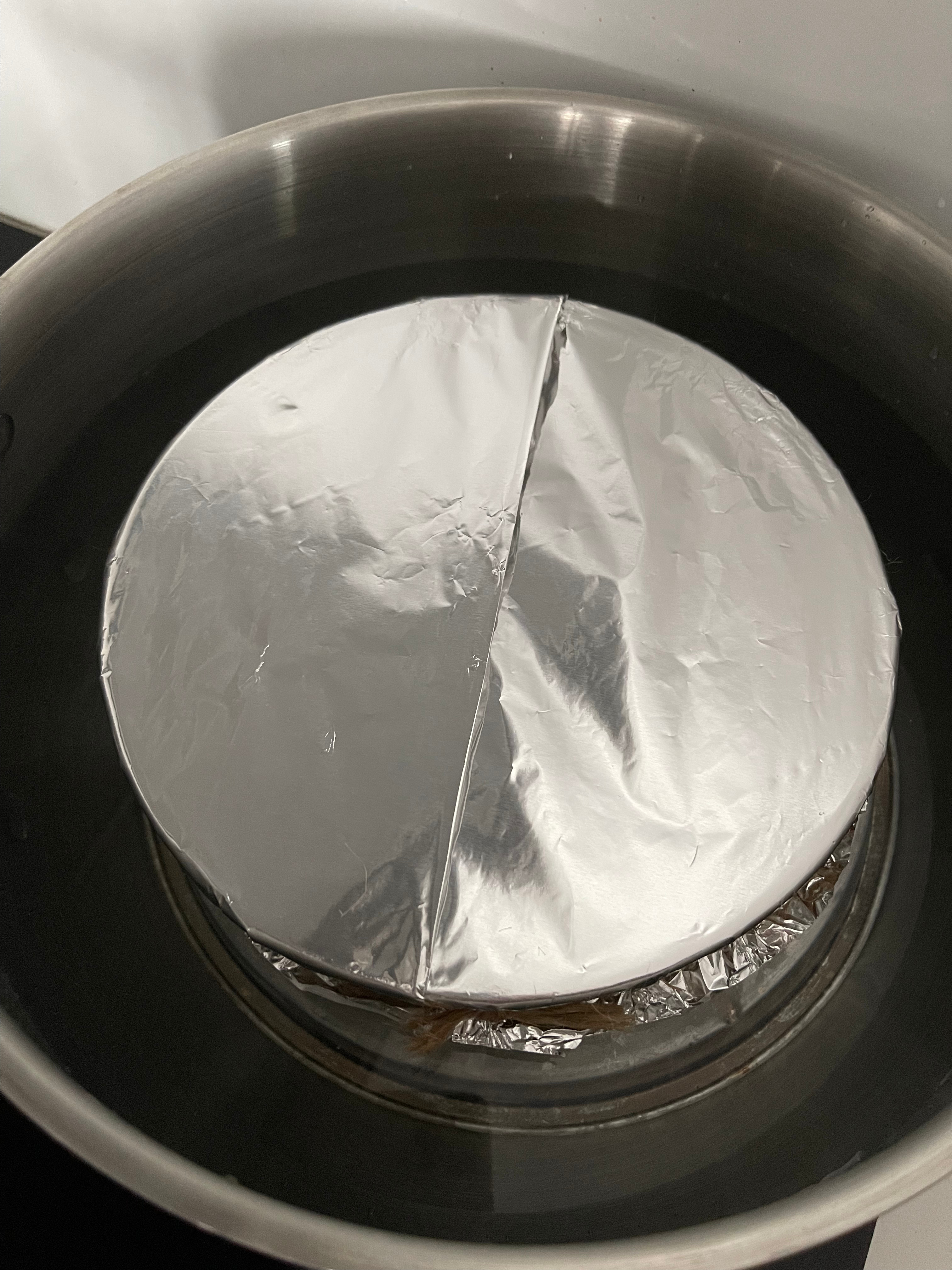 A tigela do bolo coberta com papel e folha de alumínio, com a prega central visível, dentro do tacho a cozer a vapor